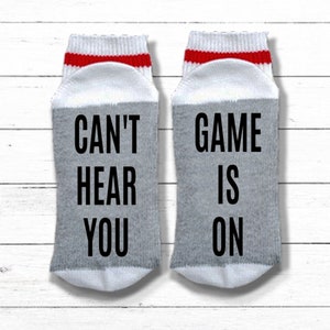 May include: A pair of gray and white socks with red stripes. One sock says "Can't Hear You" and the other says "Game Is On".