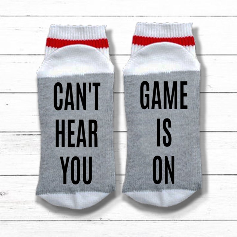 May include: A pair of gray and white socks with red stripes. One sock says "Can't Hear You" and the other says "Game Is On".