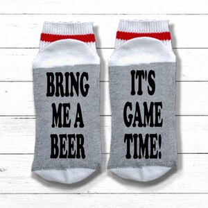 May include: A pair of gray and white socks with red stripes. One sock says "Bring me a beer" and the other says "It's game time!"