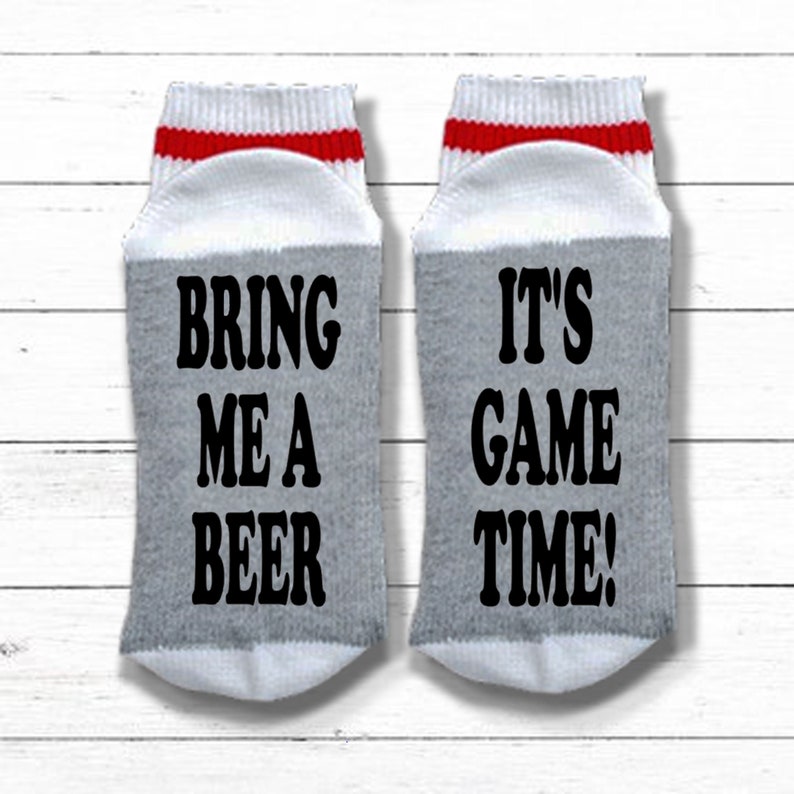 May include: A pair of gray and white socks with red stripes. One sock says "Bring me a beer" and the other says "It's game time!"