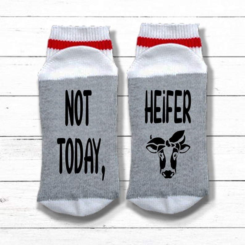 K&ouml;nnte beinhalten: Ein Paar graue und wei&szlig;e Socken mit roten Streifen. Eine Socke sagt "NOT TODAY," und die andere Socke sagt "HEIFER" mit einer Schwarzwei&szlig;-Kuh-Illustration.