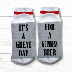 K&ouml;nnte beinhalten: Ein Paar graue und wei&szlig;e Socken mit roten Streifen. Eine Socke sagt "It's a Great Day" und die andere Socke sagt "For a Guinness Beer".