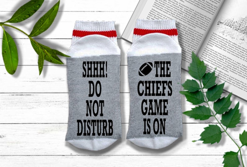 Peut inclure: Une paire de chaussettes grises avec des d&eacute;tails blancs et rouges. L'une des chaussettes porte l'inscription "SHHH! DO NOT DISTURB" en noir, et l'autre "THE CHIEFS GAME IS ON" avec un ballon de football. Les chaussettes sont sur une surface en bois blanc.