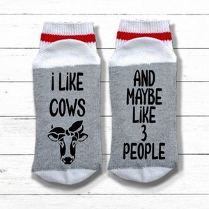 K&ouml;nnte beinhalten: Ein Paar graue und wei&szlig;e Socken mit roten und wei&szlig;en Streifen oben. Eine Socke sagt "ich mag K&uuml;he" mit einer Schwarzwei&szlig;-Kuh-Illustration. Die andere Socke sagt "UND VIELLEICHT MAG ICH 3 MENSCHEN".