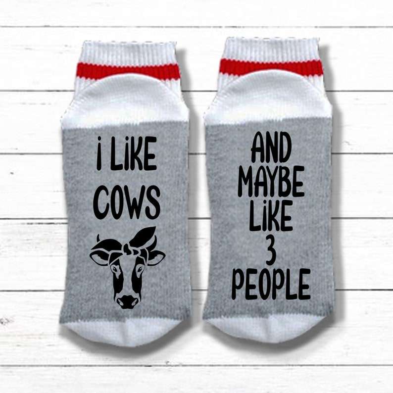 K&ouml;nnte beinhalten: Ein Paar graue und wei&szlig;e Socken mit roten und wei&szlig;en Streifen oben. Eine Socke sagt "ich mag K&uuml;he" mit einer Schwarzwei&szlig;-Kuh-Illustration. Die andere Socke sagt "UND VIELLEICHT MAG ICH 3 MENSCHEN".