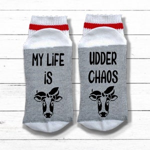 K&ouml;nnte beinhalten: Ein Paar graue und wei&szlig;e Socken mit roten Streifen. Eine Socke sagt "My life is" mit einer Kuh-Illustration. Die andere Socke sagt "Udder Chaos" mit einer Kuh-Illustration.
