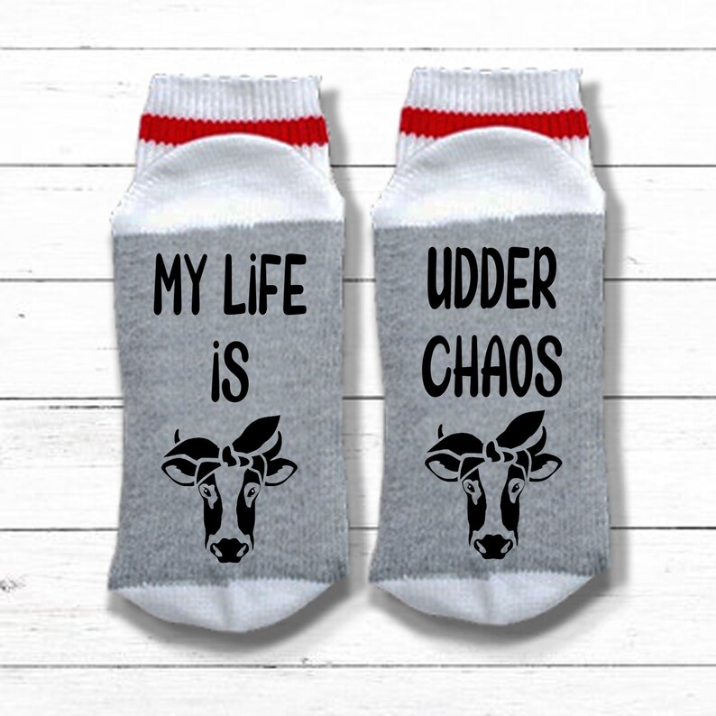 K&ouml;nnte beinhalten: Ein Paar graue und wei&szlig;e Socken mit roten Streifen. Eine Socke sagt "My life is" mit einer Kuh-Illustration. Die andere Socke sagt "Udder Chaos" mit einer Kuh-Illustration.