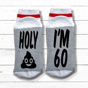 May include: A pair of white socks with red and white stripes at the top. One sock says "HOLY" with a poop emoji and the other sock says "I'M 60".