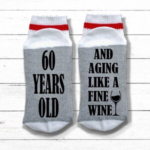 May include: A pair of gray and white socks with red stripes. One sock says "60 Years Old" and the other sock says "And Aging Like A Fine Wine" with a wine glass graphic.