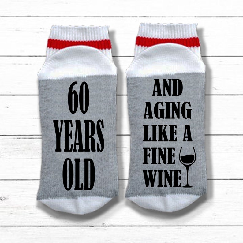 May include: A pair of gray and white socks with red stripes. One sock says "60 Years Old" and the other sock says "And Aging Like A Fine Wine" with a wine glass graphic.