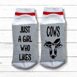 K&ouml;nnte beinhalten: Ein Paar graue und wei&szlig;e Socken mit roten Streifen. Eine Socke sagt "Just a girl who likes cows" und die andere Socke hat ein schwarz-wei&szlig;es Kuh-Motiv mit einem Bandana.