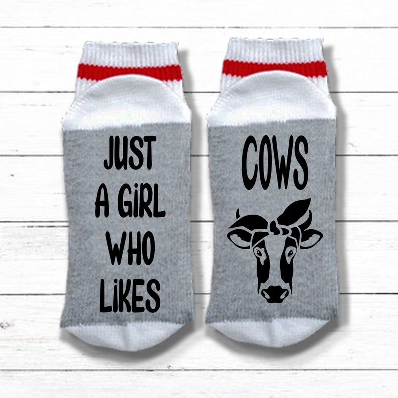 K&ouml;nnte beinhalten: Ein Paar graue und wei&szlig;e Socken mit roten Streifen. Eine Socke sagt "Just a girl who likes cows" und die andere Socke hat ein schwarz-wei&szlig;es Kuh-Motiv mit einem Bandana.