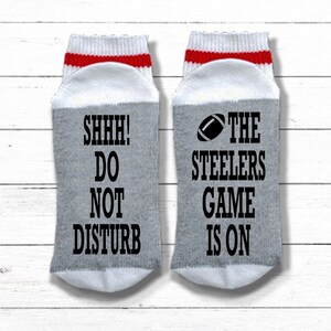 May include: A pair of gray and white socks with red stripes. One sock says "SHHH! DO NOT DISTURB" and the other says "THE STEELERS GAME IS ON" with a football graphic.