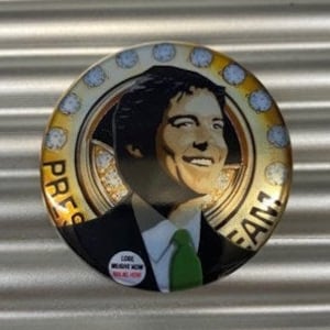 May include: A gold and black button with a portrait of a man wearing a suit and green tie. The button has a diamond-like pattern around the edge and the words "Press Team" in a circle around the portrait.