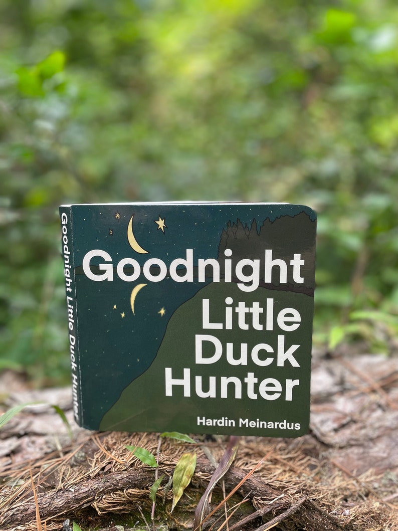 May include: A green book titled "Goodnight Little Duck Hunter" by Hardin Meinardus. The cover features a crescent moon and stars against a dark green background.