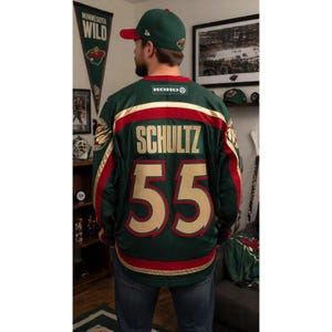 May include: A green and red hockey jersey with the name "SCHULTZ" and the number "55" in gold. The jersey has gold and red stripes and the "KOHO" logo. A green baseball cap with a red brim is also visible.