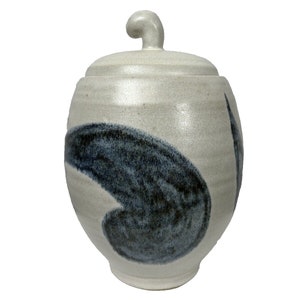 Könnte beinhalten: Keramiktopf mit Deckel, hellbeige mit dunkelblauem, abstraktem Design. Der Topf hat eine runde Form und einen geschwungenen Griff auf dem Deckel. Das Design erinnert an einen Pinselstrich.