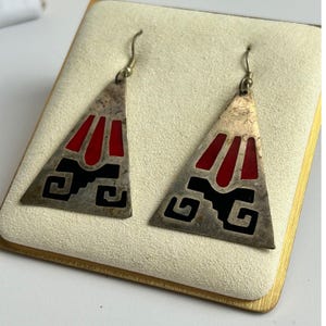 May include: A pair of triangular drop earrings featuring a tarnished silver-tone metal finish. Each earring displays inlaid red and black enamel sections arranged in a geometric pattern, suspended from simple silver-tone metal ear wire hooks.