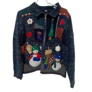May include: A blue zip-up sweater with Christmas-themed appliqués. The sweater features a patchwork design with snowmen, Christmas trees, and snowflakes. The collar is a darker blue, and the sweater has a zipper closure.