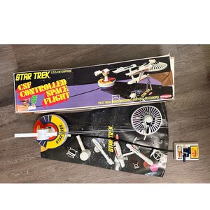 Może przedstawiać: Zestaw zabawek vintage Star Trek U.S.S. Enterprise Controlled Space Flight firmy Remco. Zawiera biały model statku kosmicznego, okrągłą żółto-niebieską bazę sterującą, ręczny kontroler oraz czarną tablicę ekspozycyjną z grafikami kosmosu.