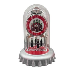 May include: A Coca-Cola branded table clock encased in a clear glass dome with a silver bottle cap base. The clock face features a red background with illustrations of glass soda bottles and black hands. Miniature soda bottles rotate below.