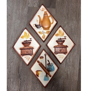 May include: Four diamond-shaped wall plaques featuring raised relief designs. One depicts a brown teapot and cup, two show brown coffee grinders, and one displays a rolling pin, a loaf of bread, and a blue jar with the letter F printed on it.
