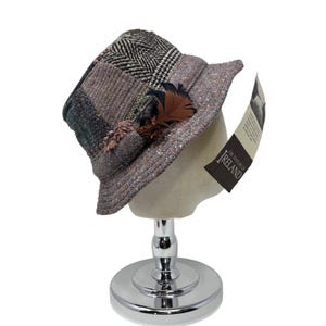 May include: A tweed patchwork hat with a brown feather detail. The hat has a mix of textures and patterns, including herringbone and solid fabrics. The hat is displayed on a mannequin head.