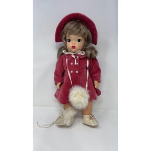 May include: A vintage doll wearing a red velvet coat and matching hat. The doll has brown hair and is wearing white shoes and fur-trimmed boots. A white pom-pom bag hangs from the coat. The doll has a classic, nostalgic appearance.