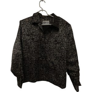 Puede incluir: Una camisa abotonada con estampado en blanco y negro con dos bolsillos en el pecho y un cuello. La camisa está colgada de una percha blanca. El estampado es un diseño similar a una cuadrícula.