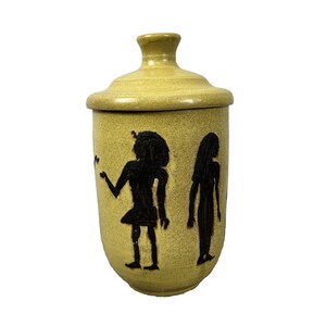 Könnte beinhalten: Ein gelbes Keramikgefäß mit Deckel, verziert mit schwarzen Silhouetten. Das Gefäß hat eine zylindrische Form mit leicht abgerundetem Boden. Der gelbe Deckel passt gut. Die Figuren erinnern an altägyptische Kunst.