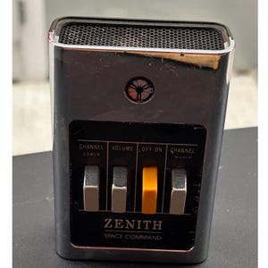 May include: A vintage Zenith Space Command television remote control with a chrome-finished rectangular body and a black control panel. It features three grey buttons and one central orange button, with labels for channel, volume, and off-on functions.
