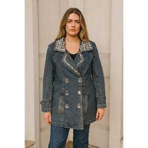 Könnte beinhalten: Eine blaue Jeansjacke mit doppelreihigem Design. Der Kragen und die Manschetten sind mit silberfarbenen Nieten verziert. Die Jacke hat große dekorative Knöpfe. Die Jacke wird mit einer blauen Jeans getragen.