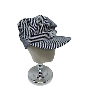 May include: A blue and white striped cap with a short brim. The cap has a rounded crown and a small rectangular patch with the text "KEY Imperial". The cap is displayed on a mannequin head with a silver stand.
