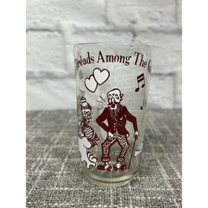 Puede incluir: Vaso transparente con una ilustración marrón de dos figuras, notas musicales y corazones. El texto "Threads Among The" está impreso en la parte superior. Las figuras son un hombre con bastón y una mujer. El vaso está sobre una superficie gris.