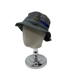 May include: A patchwork bucket hat in shades of grey, blue, and brown, displayed on a mannequin head. The hat features a blue band and decorative elements. The hat is made of a textured fabric.