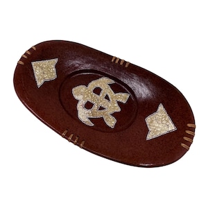 Könnte beinhalten: Ovalförmige, braune Dekoschale mit einem Schildkrötenmuster in der Mitte. Die Schale hat eine strukturierte Oberfläche und ist mit hellfarbigen, geometrischen Akzenten verziert. Die Ränder sind mit einem helleren braunen Faden vernäht.