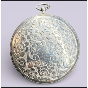 May include: A silver pocket watch with an ornate floral design. The watch has a circular shape with a loop at the top. The surface is engraved with detailed flowers and swirling patterns. The words "GERMAN SILVER" are visible.