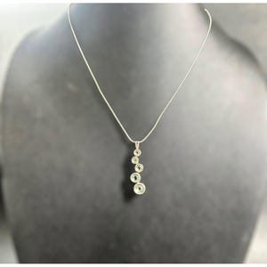 May include: A silver-toned necklace featuring a delicate chain and a vertical pendant composed of five circular settings, each containing a small, light-colored stone arranged in a descending size order from top to bottom.