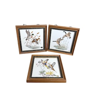 May include: Set of three framed ceramic tiles featuring detailed illustrations of various birds in flight and in their natural habitat. Each tile is set in a wooden frame with a dark trim. The artwork depicts ducks, geese, and pheasants.