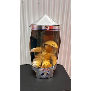 Puede incluir: Muñeca astronauta Cabbage Patch Kids en una vitrina de plástico transparente. La muñeca lleva un traje y casco de astronauta amarillos. La vitrina tiene una tapa plateada y una base con las palabras "Cabbage Patch Kids Astronaut Out of This World."
