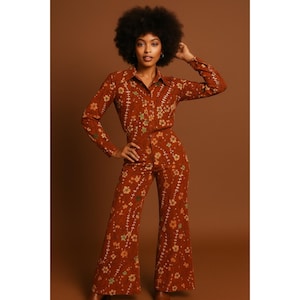 May include: A rust-coloured two-piece outfit, comprising a button-down shirt and flared trousers. The ensemble is decorated with a floral pattern in yellow, green, and white. The outfit has a retro, 1970s style.