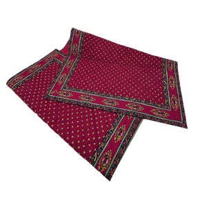 Può includere: Due tovagliette color bordeaux con un motivo ripetuto di piccole croci gialle. I bordi presentano un design ornato con accenti neri, oro e rosa. Le tovagliette sono piegate e sovrapposte.