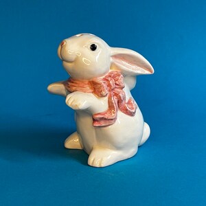 May include: A white ceramic rabbit figurine with a pink bow. The rabbit is standing on all four legs and has its head tilted to the side.