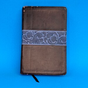 Puede incluir: Un diario de cuero marrón encuadernado con una banda de patrón azul. El diario tiene un marcador de cinta negro.