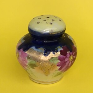 May include: A small, blue and white porcelain salt shaker with a floral design and gold accents. The shaker has a round body and a domed lid with holes for dispensing salt.