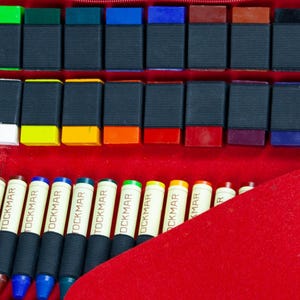 May include: A set of 24 colorful wax crayons in a red case. The crayons are arranged in rows and labeled "TOCKMAR".
