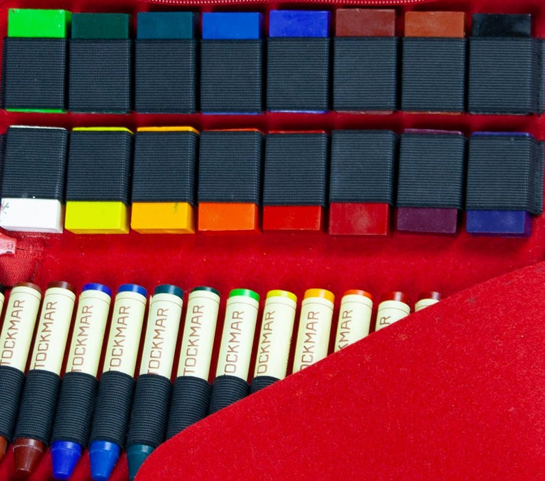 May include: A set of 24 colorful wax crayons in a red case. The crayons are arranged in rows and labeled "TOCKMAR".