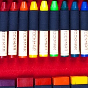 May include: A set of 12 crayons in a variety of colors, including red, orange, yellow, green, blue, purple, and brown. The crayons are arranged in a row and are labeled "TOCKMAR" on the side.