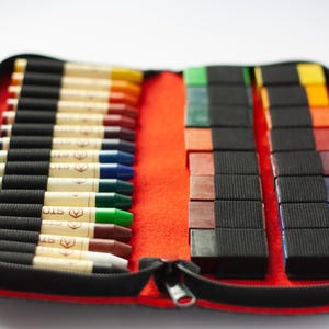 May include: A red zippered case with a black lining holds a set of crayons. The crayons are arranged in two rows, with the longer crayons on the left and the shorter crayons on the right. The case is open, revealing the crayons inside.