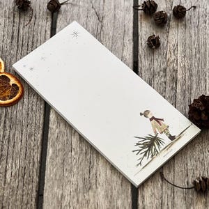 Könnte beinhalten: Ein rechteckiger weißer Notizblock mit einer Winterlandschaftsillustration. Die Illustration zeigt eine Person, die einen Weihnachtsbaum trägt. Der Notizblock ist mit kleinen Schneeflocken verziert. Getrocknete Orangenscheiben und Tannenzapfen sind verstreut.